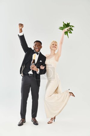 Photo for A beautiful blonde bride in a wedding dress and an African American groom holding flowers in a studio on a grey background. - Royalty Free Image