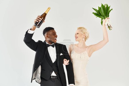 Eine schöne blonde Braut im Brautkleid und ein afroamerikanischer Bräutigam im Smoking, der eine Flasche Champagner in der Hand hält, um anzustoßen.
