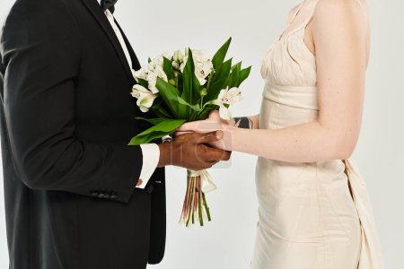 An African American groom in a tuxedo holds a bouquet of flowers next to a beautiful blonde bride in her wedding dress.