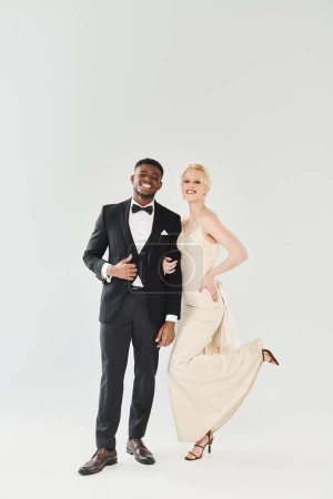 Elegant man in tuxedo stands next to a beautiful woman in a white dress in a studio setting with a grey background.