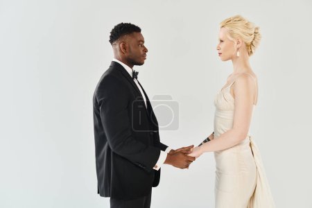 Une belle mariée blonde dans une robe de mariée et un marié afro-américain debout dans un studio sur un fond gris.