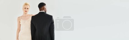 Foto de Una hermosa novia rubia con un vestido de novia y un novio afroamericano de pie lado a lado en un estudio sobre un fondo gris. - Imagen libre de derechos
