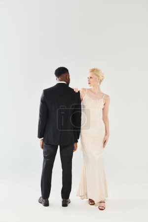 Une belle mariée blonde dans une robe de mariée et un marié afro-américain se tiennent côte à côte dans un studio sur un fond gris.