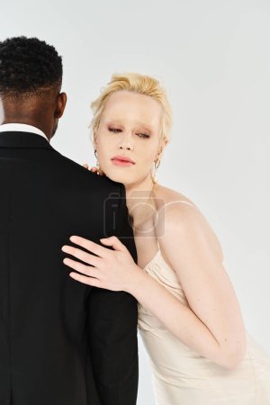 Una hermosa novia rubia con un vestido blanco y un novio afroamericano con un esmoquin se paran juntos en un fondo gris de estudio.