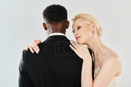 A beautiful blonde bride in a wedding dress and an African American groom embracing each other passionately in a studio on a grey background.