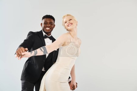 A beautiful blonde bride in a wedding dress stands beside an African American groom in a sharp tuxedo on a grey background.
