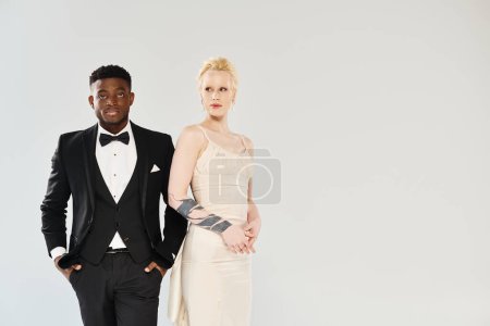 An African American man in a tuxedo stands next to a beautiful blonde woman in a white dress in a studio against a grey background.