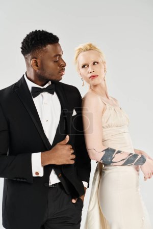A stunning blonde bride in white wedding dress and an African American groom in tuxedo stand together, radiating style and grace.
