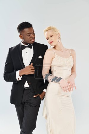 An African American groom in a tuxedo and a beautiful blonde bride in a wedding dress standing together in a studio on a grey background.