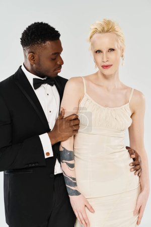 African American groom and beautiful blonde bride, dressed in tuxedo and gown, stand gracefully in a studio against a grey background.