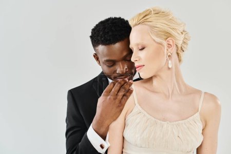 Foto de Una hermosa novia rubia en un vestido de novia está junto a su novio afroamericano en un estudio sobre un fondo gris. - Imagen libre de derechos