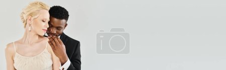 Téléchargez les photos : Une belle mariée blonde dans une robe de mariée se tient à côté d'un marié afro-américain dans un studio sur un fond gris. - en image libre de droit