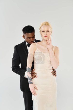Une belle mariée blonde dans une robe de mariée blanche debout à côté d'un marié afro-américain dans un studio sur un fond gris.