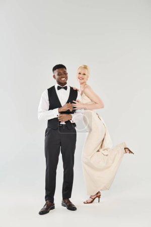 Téléchargez les photos : Une belle mariée blonde dans une robe de mariée et un marié afro-américain dans une pose de smoking ensemble dans un studio sur fond gris. - en image libre de droit