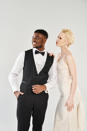 Eine schöne blonde Braut im weißen Brautkleid steht neben ihrem afroamerikanischen Bräutigam im Smoking und strahlt Eleganz und Liebe aus.