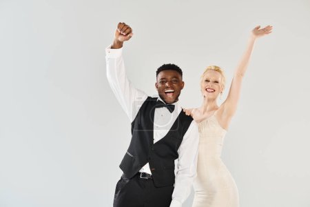 Une belle mariée blonde dans une robe de mariée et un marié afro-américain posent élégamment dans un studio sur un fond gris.