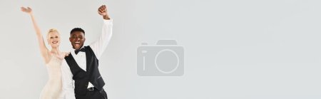 Foto de Una hermosa novia rubia con un vestido de novia y un novio afroamericano saltan alegremente al aire contra un fondo gris del estudio. - Imagen libre de derechos