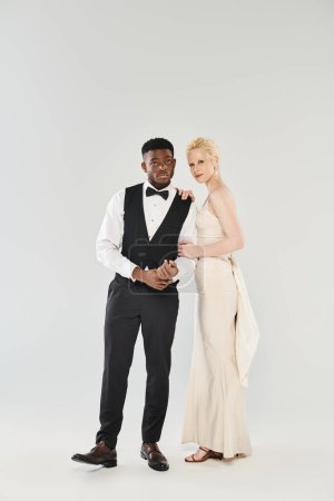 An African American groom in a tuxedo and a beautiful blonde bride in a flowing wedding dress pose elegantly in a studio setting.