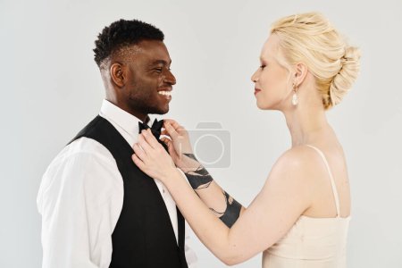 una hermosa novia rubia ayudando a su novio afroamericano a ponerse la corbata en un estudio sobre un fondo gris.