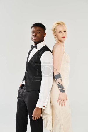 Une belle mariée blonde dans une robe de mariée et un marié afro-américain debout ensemble dans un studio sur un fond gris.