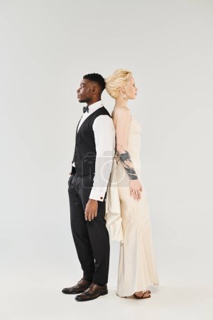 Eine schöne blonde Braut im Brautkleid und ein afroamerikanischer Bräutigam stehen nebeneinander in einem Studio vor grauem Hintergrund..