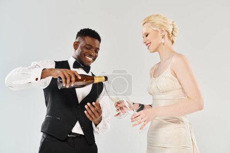 Un hombre con esmoquin vierte champán en una mano de mujer, mientras celebran en un estudio con una hermosa novia rubia en un vestido de novia y un novio afroamericano sobre un fondo gris.