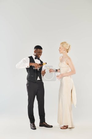 Eine schöne blonde Braut im Brautkleid und ein afroamerikanischer Bräutigam stehen nebeneinander in einem Studio auf grauem Hintergrund.