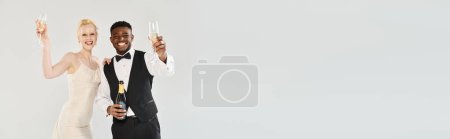 Téléchargez les photos : Une belle mariée blonde en robe de mariée et un marié afro-américain tenant des flûtes à champagne dans un studio sur fond gris. - en image libre de droit