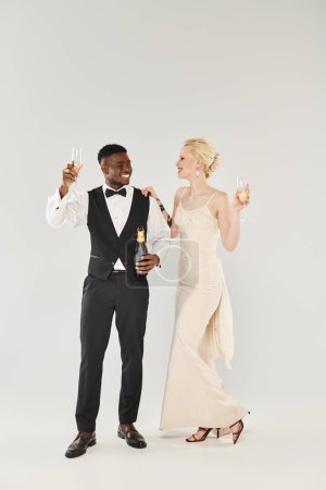 Eine schöne blonde Braut im Hochzeitskleid und ein afroamerikanischer Bräutigam mit Champagnerflöten in einem Studio vor grauem Hintergrund.