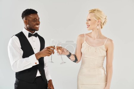 Hermosa novia rubia en vestido de novia y novio afroamericano sosteniendo copas de champán en un estudio sobre un fondo gris.