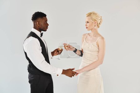 Foto de Novia rubia en vestido de novia y novio afroamericano tintinean copas de champán en un momento romántico sobre un fondo gris. - Imagen libre de derechos