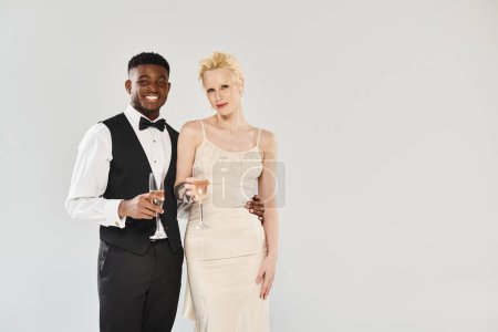 A beautiful blonde bride in a wedding dress and an African American groom in a tuxedo pose elegantly in a studio on a grey background.