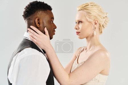 A beautiful blonde bride in a wedding dress and African American groom standing together in a studio on a grey background.