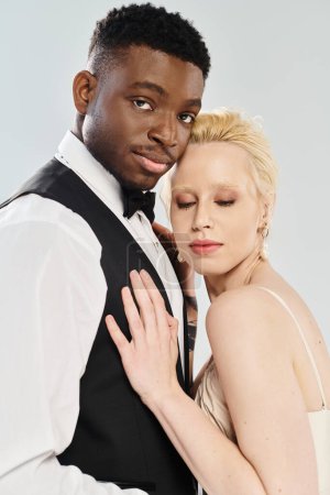 Belle mariée blonde en robe de mariée et marié afro-américain posant ensemble en studio sur fond gris.