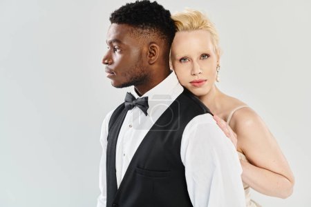 Eine schöne blonde Braut im Hochzeitskleid steht neben einem afroamerikanischen Bräutigam im Smoking vor einer grauen Studiokulisse.