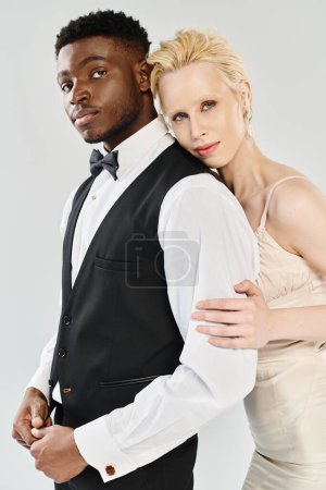 Una hermosa novia rubia con un vestido de novia y un novio afroamericano con esmoquin en un estudio sobre un fondo gris.