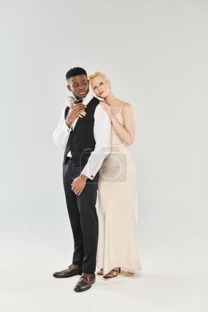 Téléchargez les photos : Une belle mariée blonde dans une robe de mariée et un marié afro-américain se tiennent côte à côte dans un studio sur un fond gris. - en image libre de droit