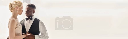 Photo for A beautiful blonde bride in a wedding dress and an African American groom stand next to each other in a studio against a grey background. - Royalty Free Image