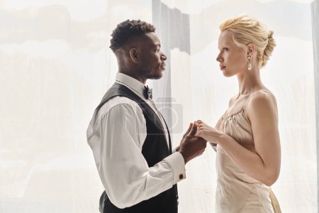 Una hermosa novia rubia con un vestido de novia y un novio afroamericano de pie juntos en un estudio, sobre un fondo gris.