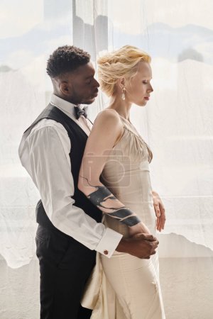 Eine schöne blonde Braut im Brautkleid und ein afroamerikanischer Bräutigam stehen nebeneinander in einem Studio vor grauem Hintergrund.