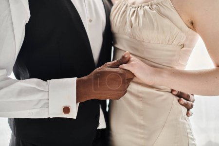 Una hermosa novia rubia con un vestido de novia y un novio afroamericano de pie uno al lado del otro en un estudio, sobre un fondo gris.