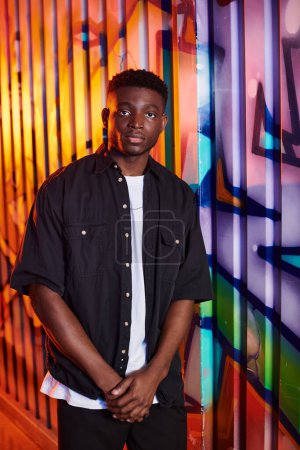 Photo for An African American man stands confidently in front of a lively, graffiti-covered wall in an urban setting. - Royalty Free Image