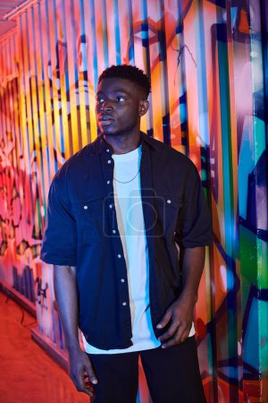 Photo for An African American man stands confidently in front of a colorful graffiti-covered wall on an urban street. - Royalty Free Image