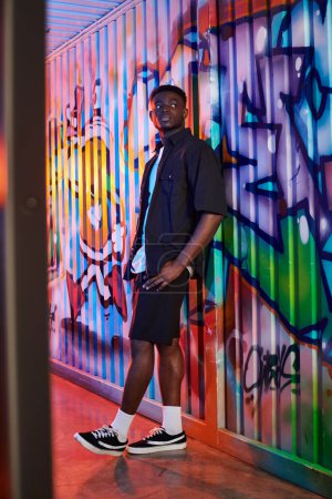Un hombre afroamericano se para con confianza frente a una pared colorida, creando un contraste visualmente llamativo en un entorno urbano.