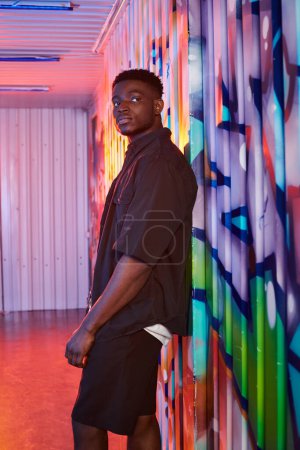 Photo for An African American man stands confidently in front of a colorful wall, showcasing a unique urban backdrop. - Royalty Free Image