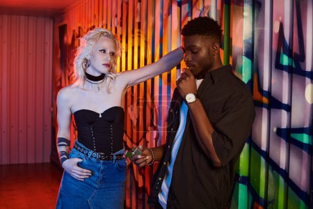 A blonde woman and African American man stand confidently in front of a colorful graffiti-covered wall in an urban setting.