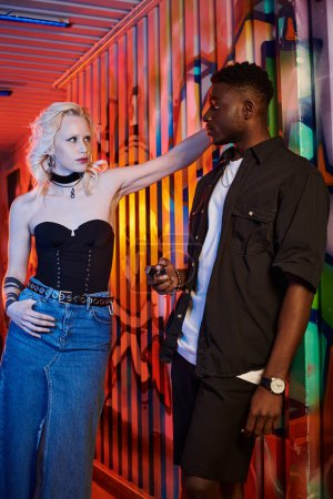 Téléchargez les photos : Une femme blonde et un homme afro-américain se tiennent côte à côte dans une rue urbaine remplie de murs recouverts de graffitis. - en image libre de droit