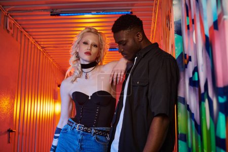 A blonde woman and an African American man stand next to each other on an urban street with graffiti-covered walls.