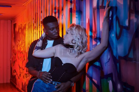 A blonde woman and African American man stand in front of a colorful wall covered in graffiti on an urban street.