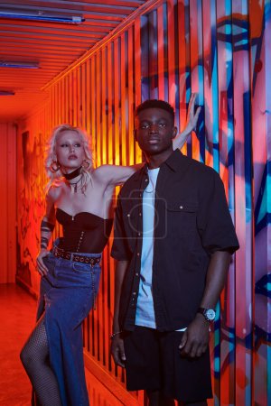 A blonde woman and an African American man stand in front of a colorful graffiti wall on an urban street.